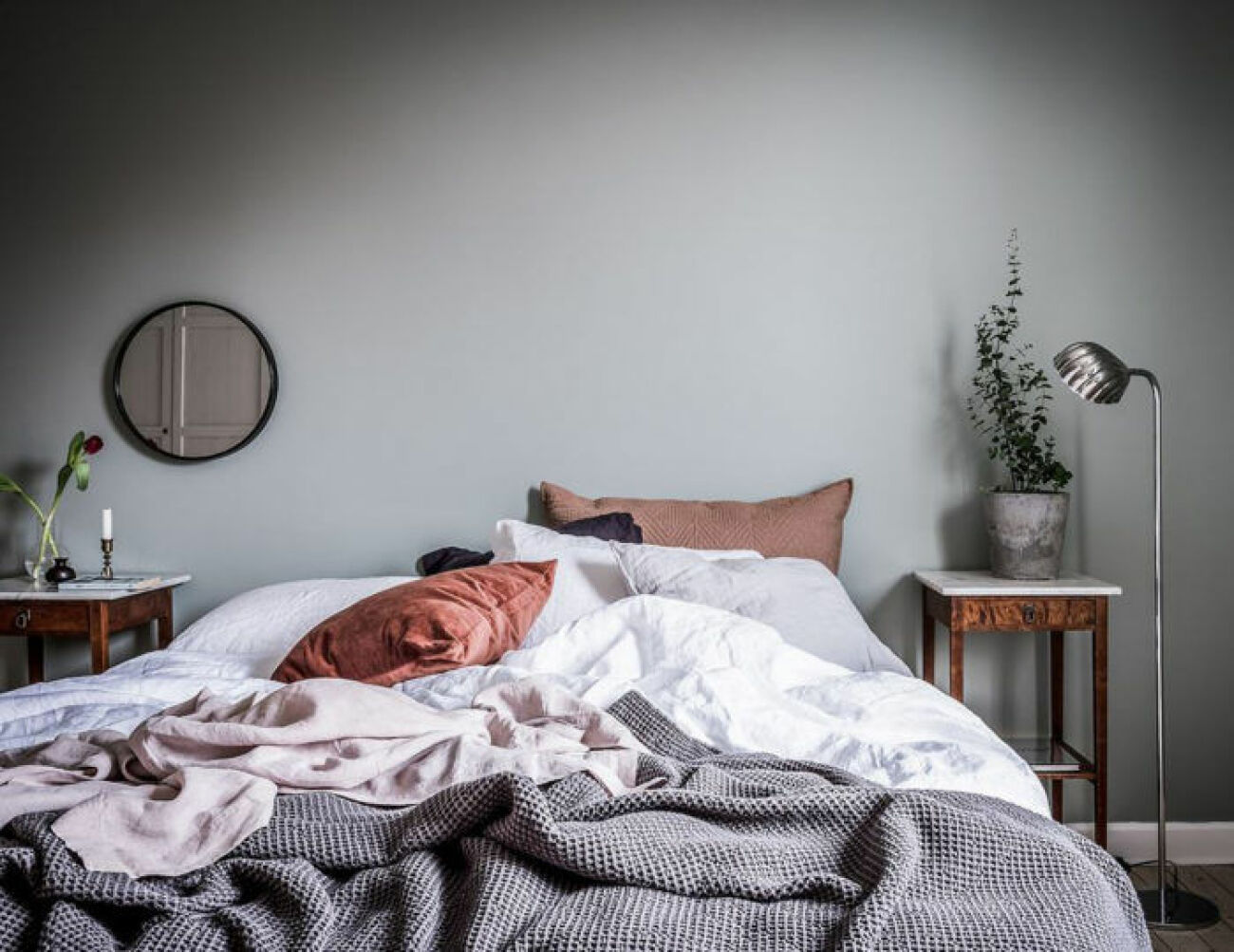 Bedroom with grey painted walls, cozy bedding and round mirror. Scandinavian decoration and ideas.