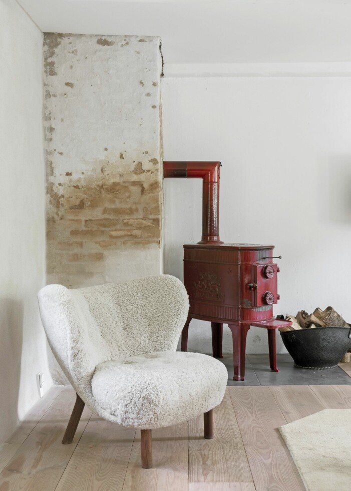 Vid den röda järnkaminen, som stod i det förfallna huset när paret köpte det, står fåtöljen Little Petra i fårskinn. Stolen är designad av Viggo Boesen 1938 och har relanserats av &Tradition. Röd järnkamin och fåtölj.