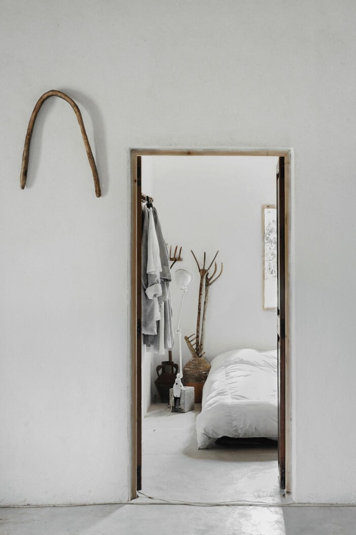 I detta vita och avskalade hem blir minsta detalj skulptural, som här i sovrummet. Kläderna på kroken är designade och uppsydda av Adriana och urnorna är Jaumes. Jieldé-lampa samt träkrattor är vintage. Inblick in i vitt avskalat sovrum genom dörröppning