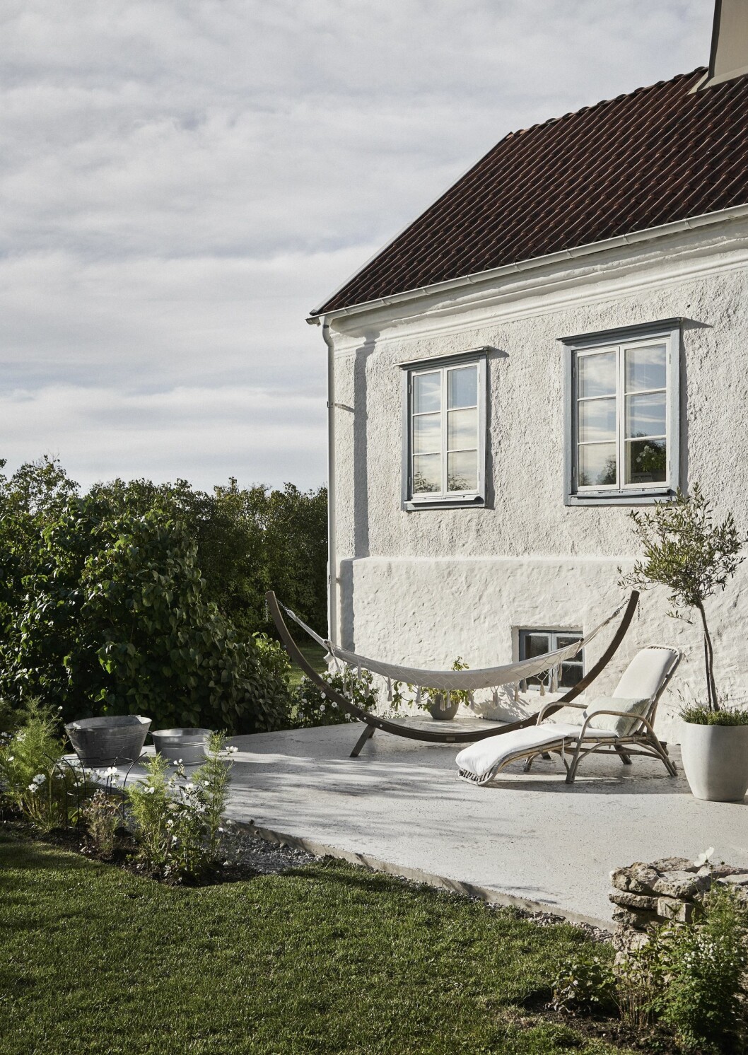Uteplatsen utanför köket har sol hela dagen och en stund på kvällen. Till höger skymtar muren som ska fortsätta byggas längs hela uteplatsen. Den putsade fasaden har de renoverat själva genom att kasta på puts som sedan strukits med kalk. Solstol i rotting från Ellos. Olivträdet kommer från Lindas syster som är florist. Stenhus med putsad fasad.