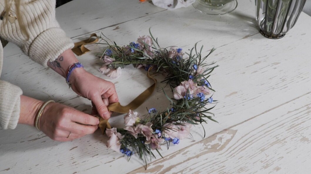 Det sista Sandra gör är att fästa ett brett sidenband i "krokarna" som hon böjt i stommen på förhand. DÅ blir midsommarkransen enkel att justera under kvällen. Steg 10 - fäst bandet i din midsommarkrans.