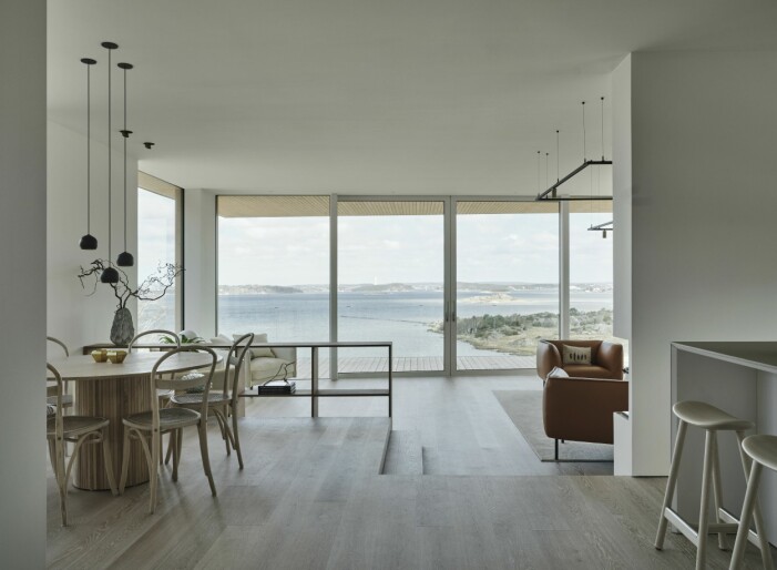 Linus ville skapa känslan av att huset svävar över havet.Två centrerade skjutdörrar leder ut till terrassen med magnifik utsikt över fjorden. Hallen leder rakt in i kök, matsal och vardagsrum i en öppen planlösning där nivåskillnaden på golvet liksom den låga bokhyllan skapar rumslighet. Vita väggar målade i NCS S0500-N tillsammans med golvet Prestige oak white sand från Tarkett skapar en enhetlig och sömlös övergång mellan rummen. Linus Fernström arkitekt